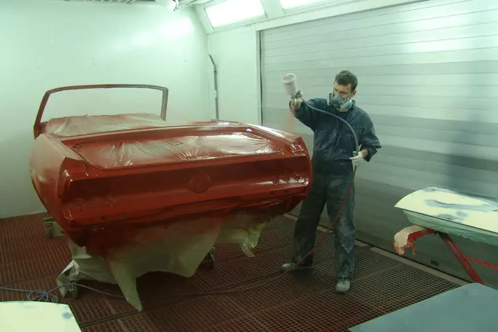 Lackierer in Schutzausrüstung trägt Basislack auf eine Mustang-Karosserie in der Lackierkabine auf